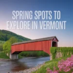 A red covered bridge spans a river surrounded by green hills and blooming pink flowers under a clear sky, inviting you to discover the best Spring Spots to Explore in Vermont.